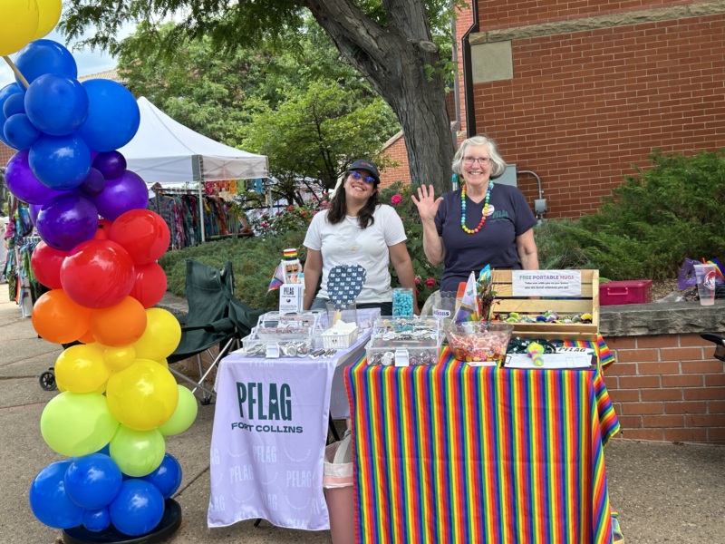 Two smiling women stand behind a PFLAG booth at a Pride event.